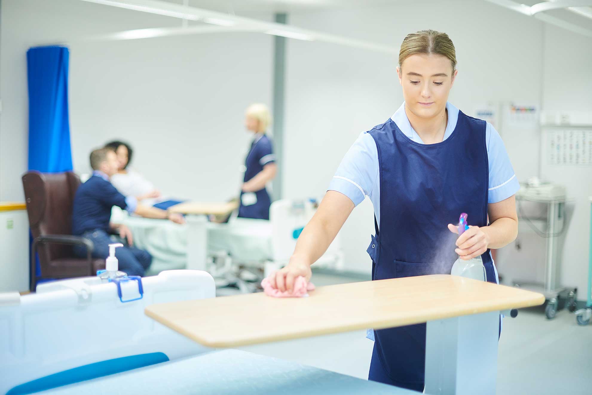 A hospital cleaner disinfecting a patient tray table on a ward